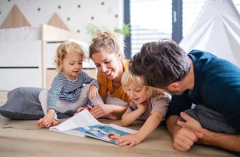 Young family with two small children indoors in bedroom reading a book.